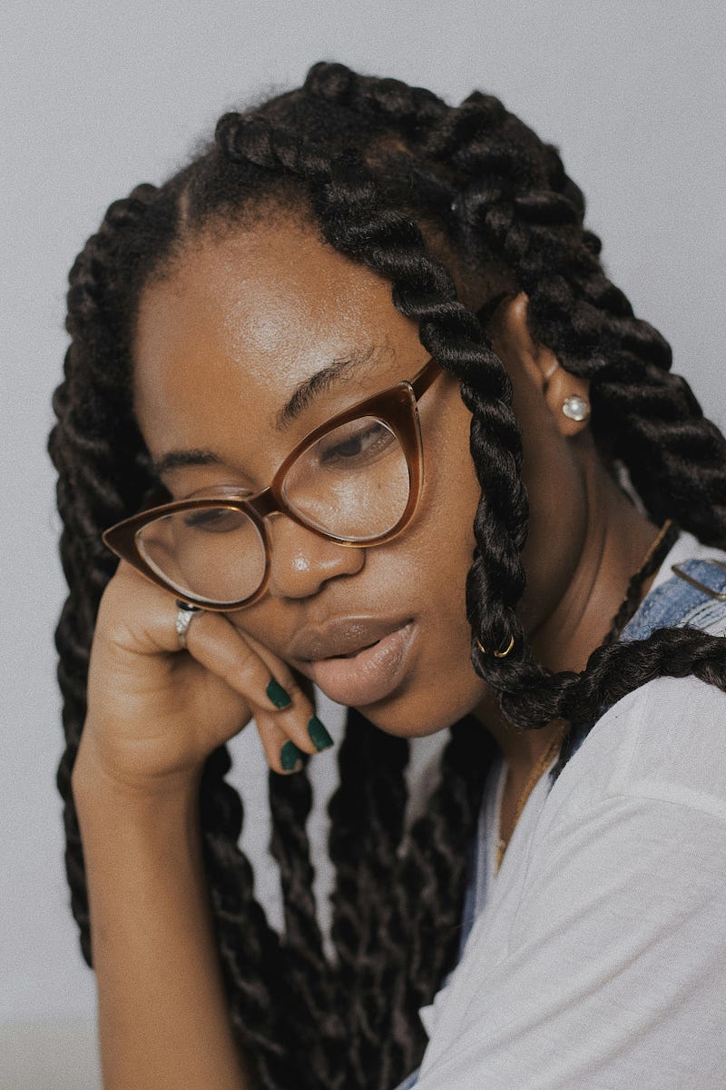 A high-achieving Black or African-American woman located in Louisiana, Texas, or Washington thinks, reflects, or contemplates about a past decision or regret. She is wearing brown glasses, and her dark hair is in twists.